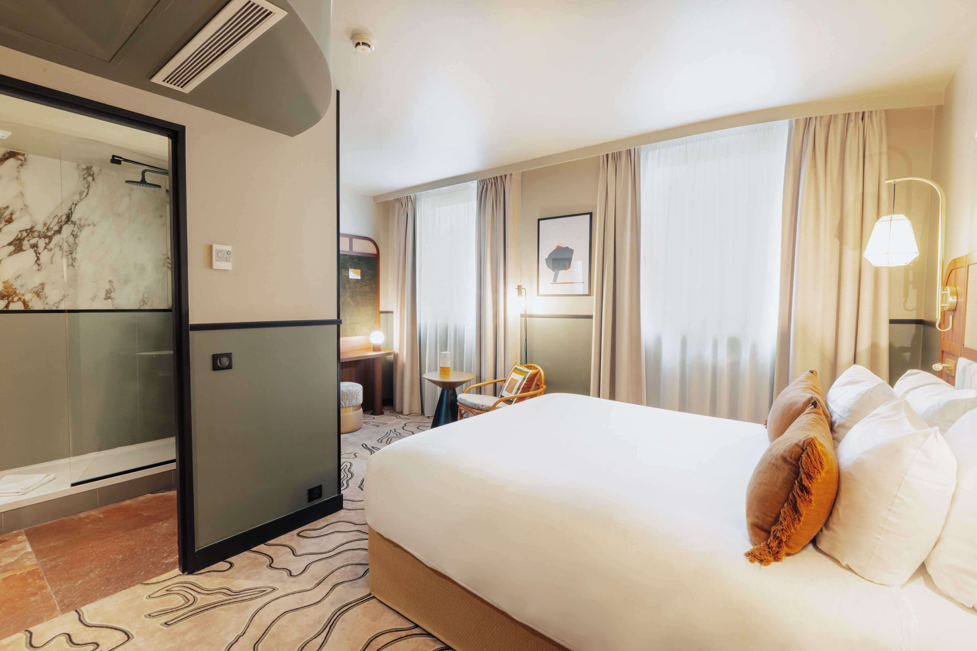 A hotel room with a large bed, a bathroom with a glass shower, a small table with a chair, and a window with curtains.