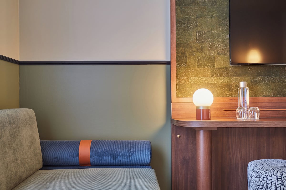 A hotel room with a sofa, a wooden side table with a lamp, and a flat-screen TV mounted on the wall.