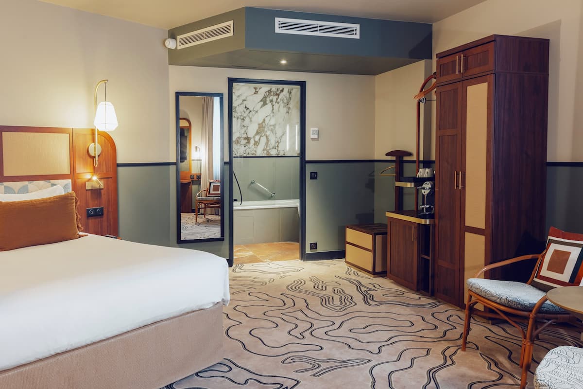 A hotel room with a bed, a wooden wardrobe, a small table, and a chair.