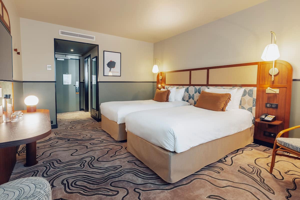 A hotel room with two beds, a desk, and a chair. There is a door leading to a bathroom and a hallway.