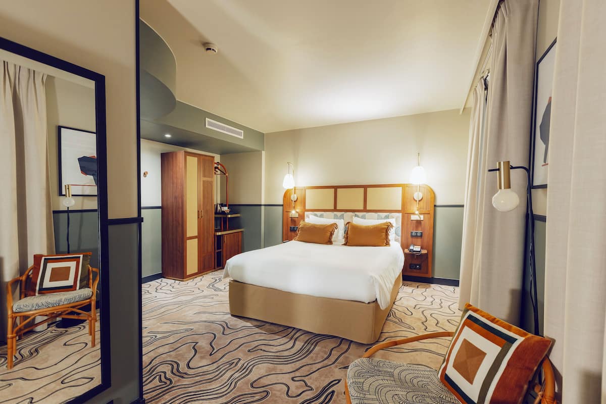 A hotel room with a large bed, a wooden wardrobe, a bedside table, and a chair.