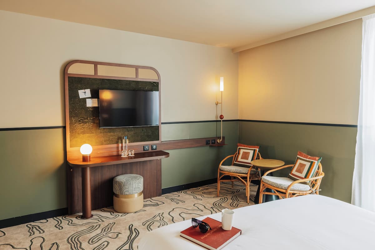 A hotel room with a bed, a desk with a flat-screen TV, a chair, a small table, and a wall-mounted light.
