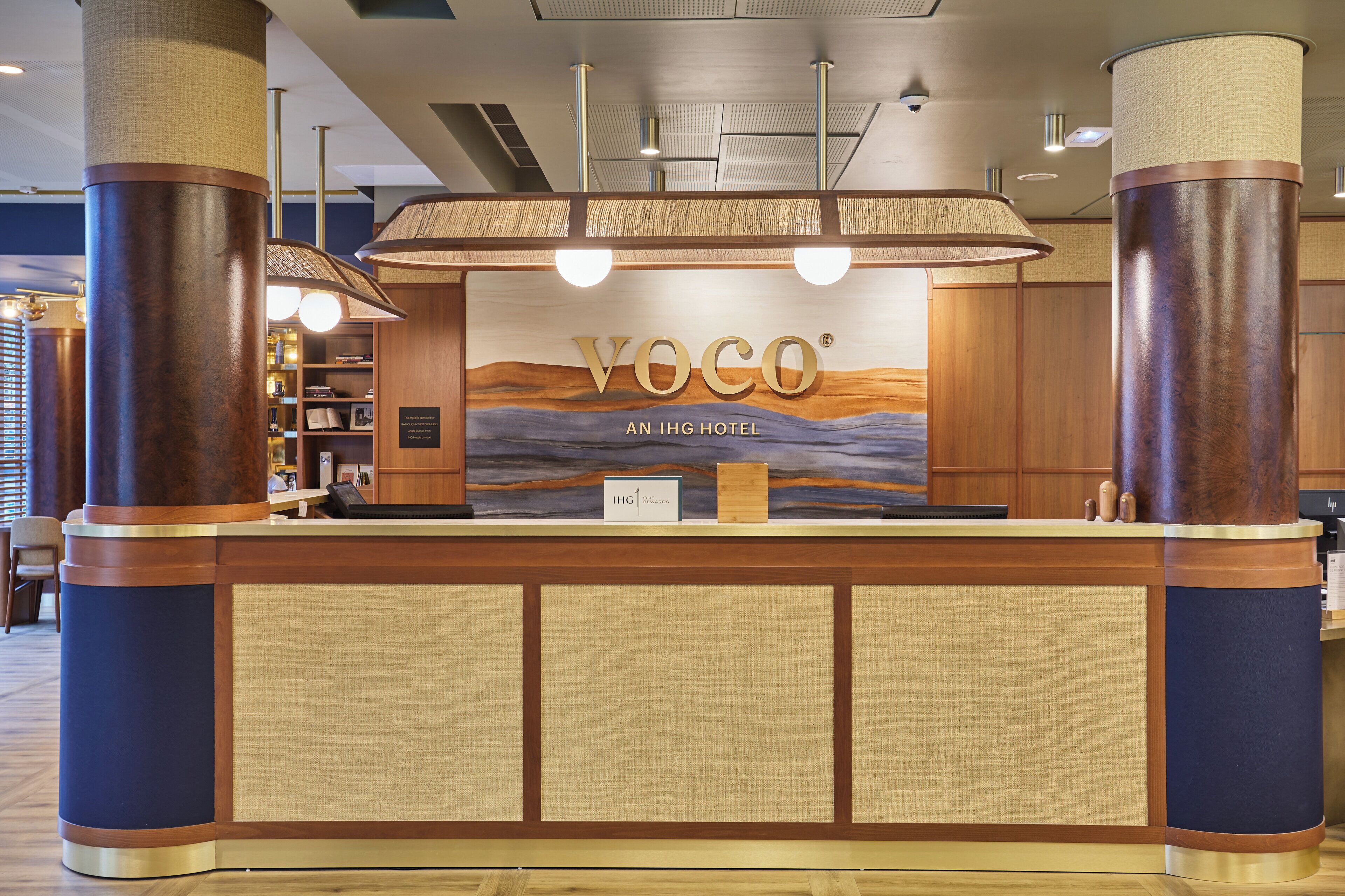 A reception desk with a wooden finish, a large sign reading "VOCO AN IHG HOTEL", and a nameplate.