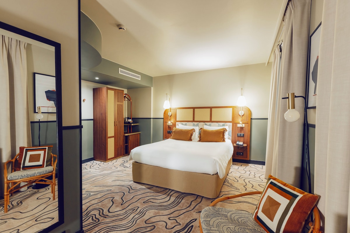 A hotel room with a large bed, a wooden wardrobe, a bedside table, and a chair.