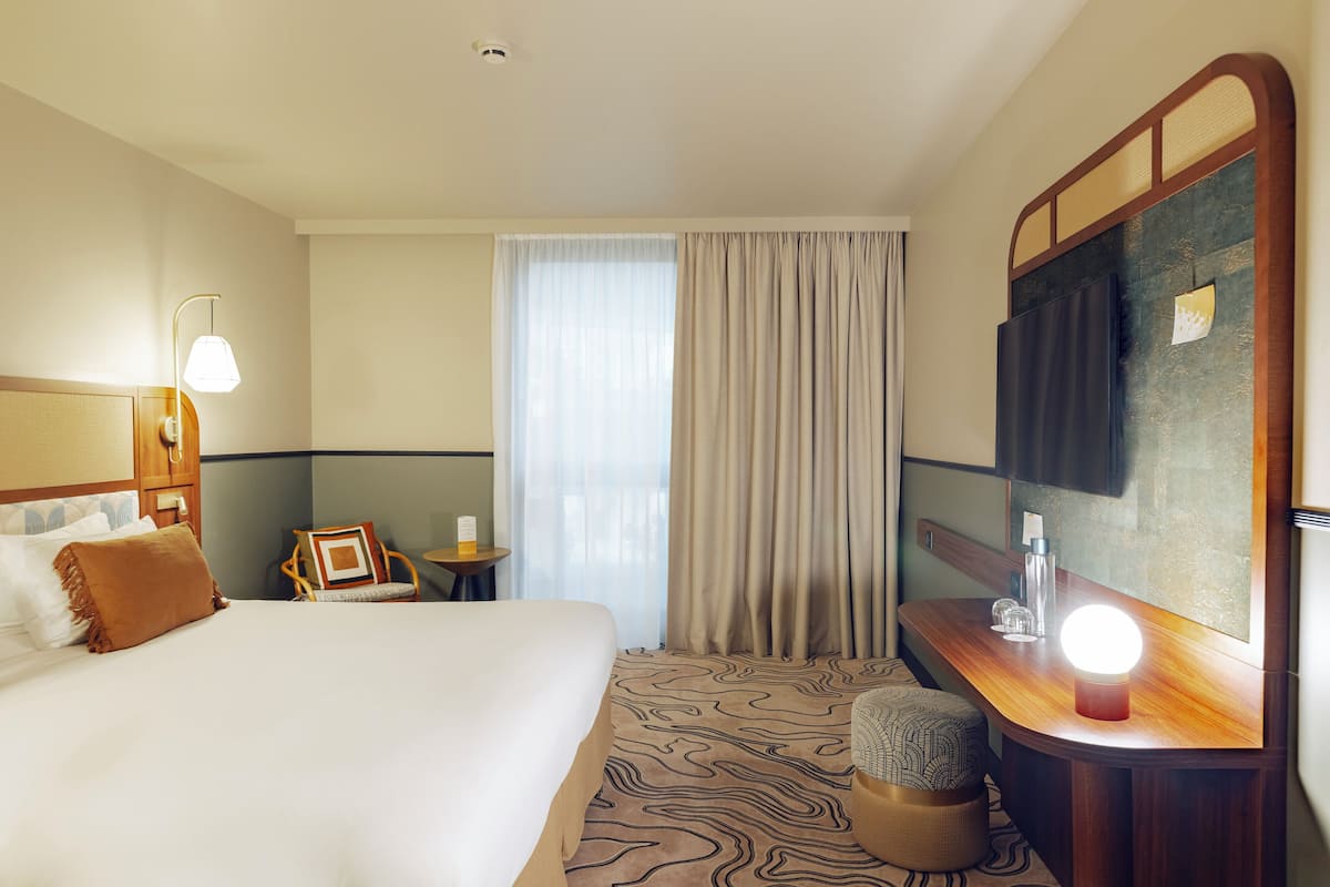 A hotel room with a large bed, a bedside table with a lamp, a small table with a chair, and a large mirror.