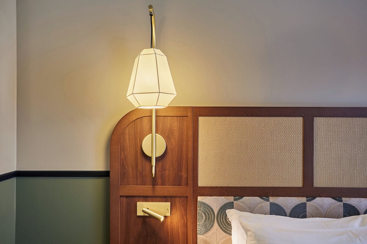 A hotel room with a wooden headboard, a wall-mounted lamp, a bedside table, and a bed with a patterned bedspread.