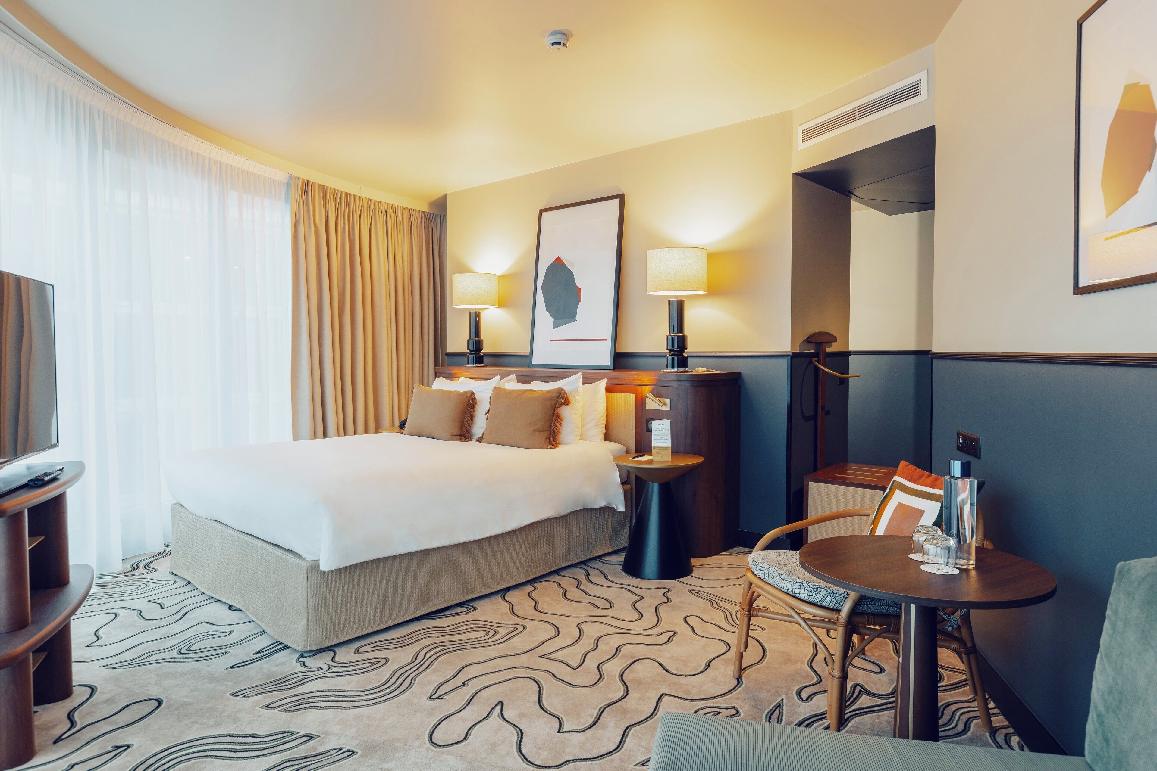 A hotel room with a large bed, two bedside lamps, a small table with a vase, and a framed picture on the wall.