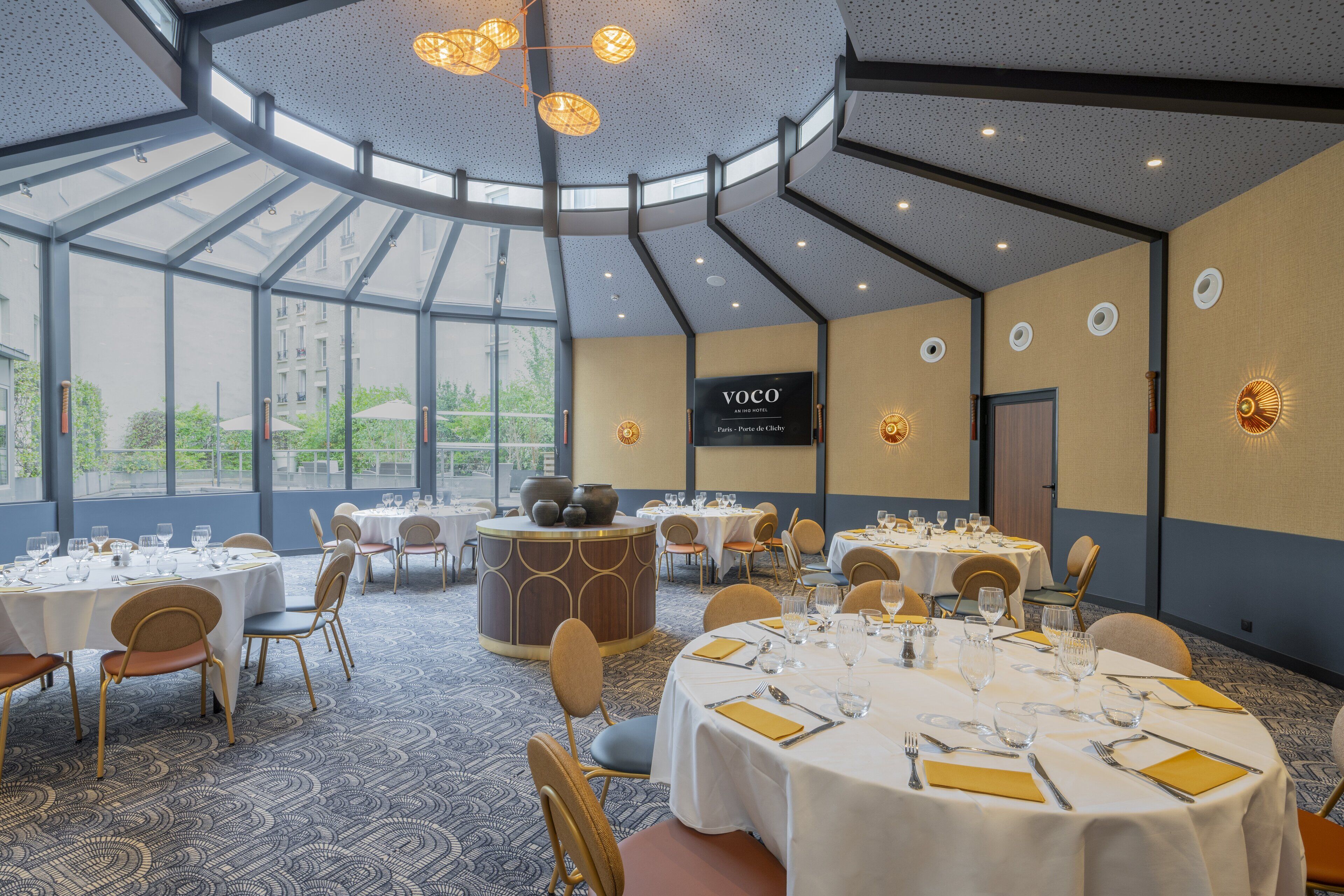 A spacious conference room with multiple tables set for dining, large windows offering a view of the city, and a modern ceiling design.