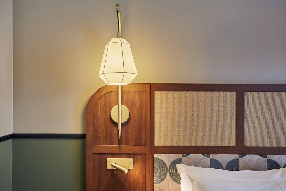 A hotel room with a wooden headboard, a wall-mounted lamp, a bedside table, and a bed with a patterned bedspread.
