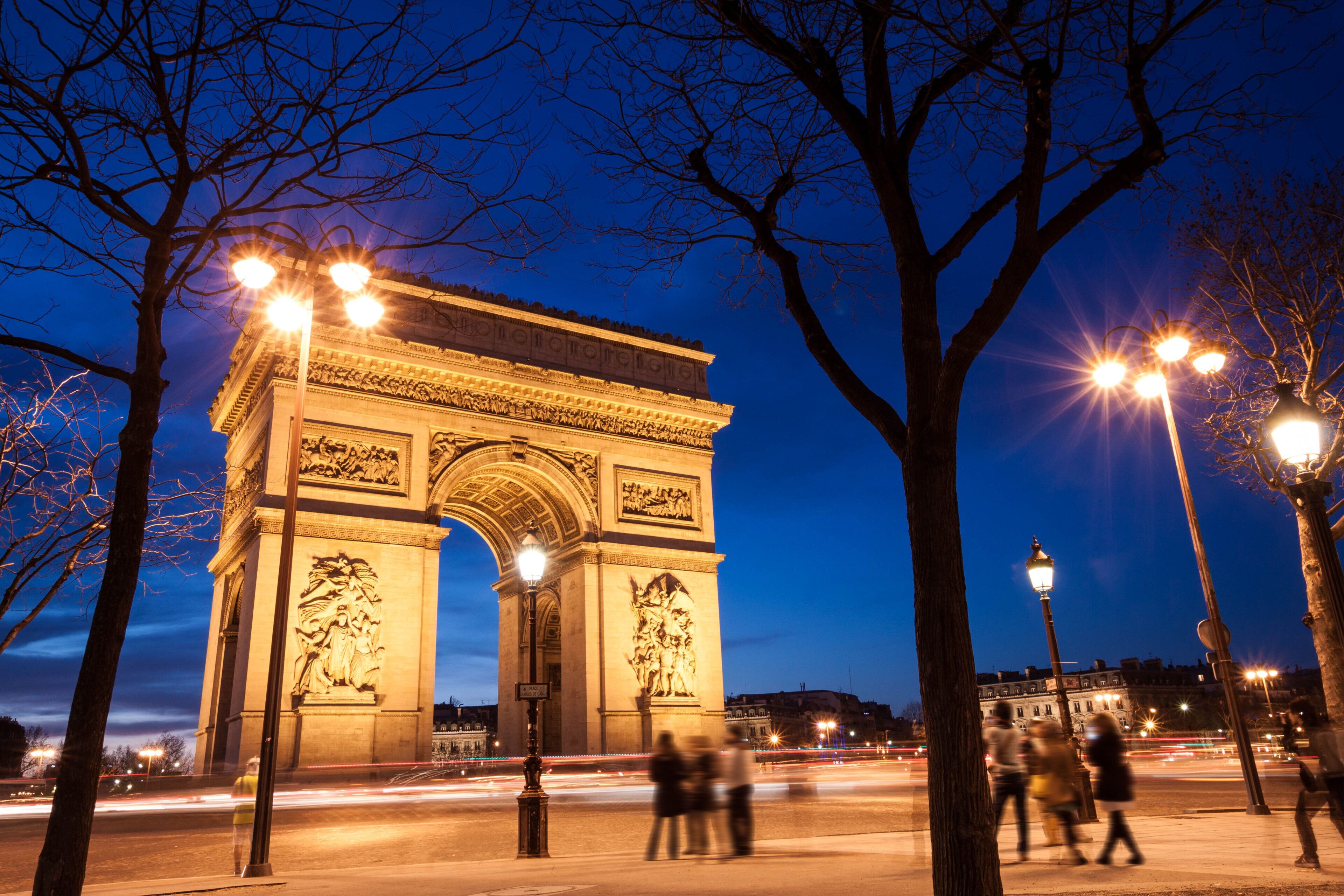 Un grand monument historique aux sculptures complexes, entouré d’arbres et de lampadaires, avec des passants en arrière-plan.