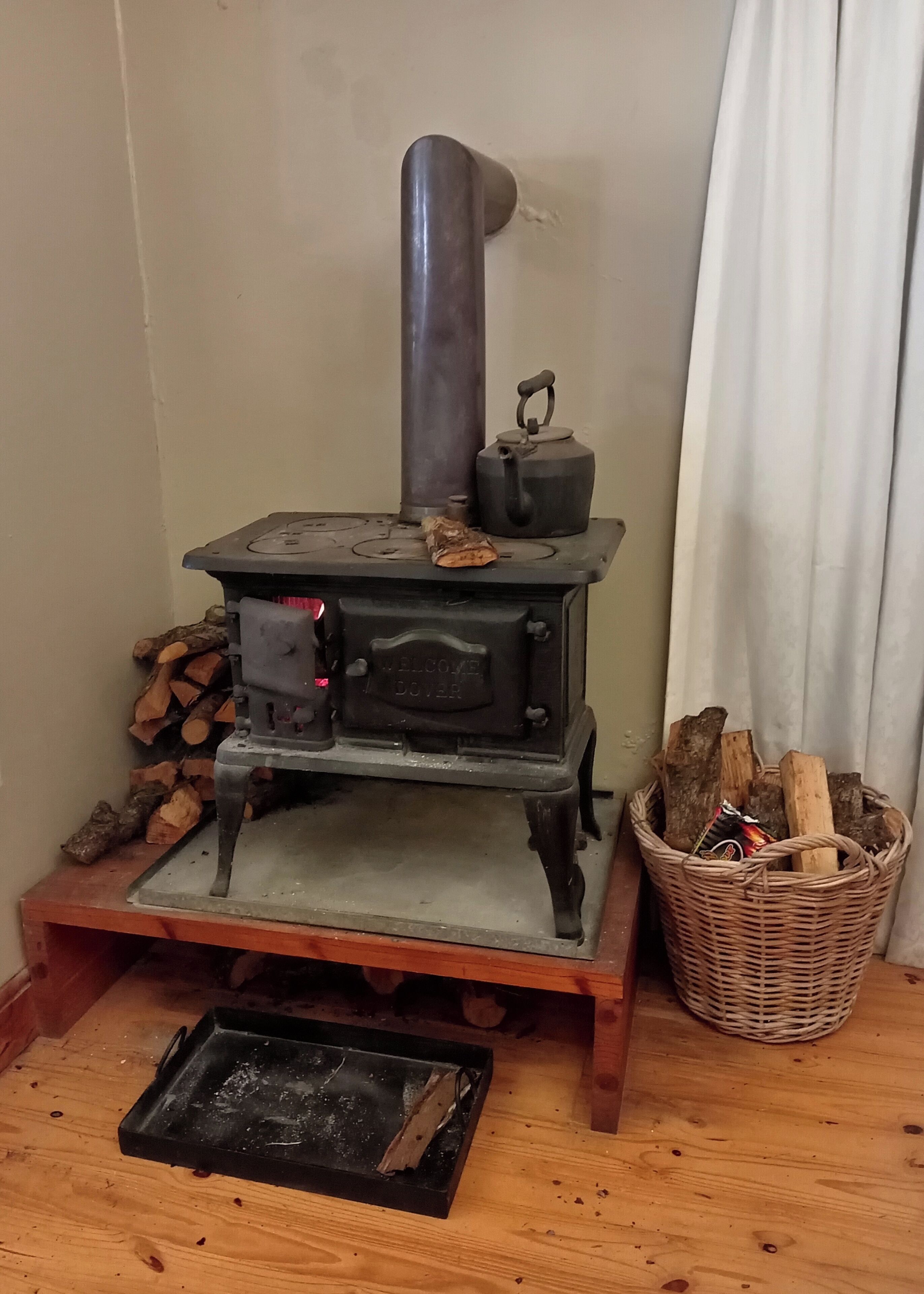 A wood-burning stove with a metal chimney, a kettle, and a basket of firewood.