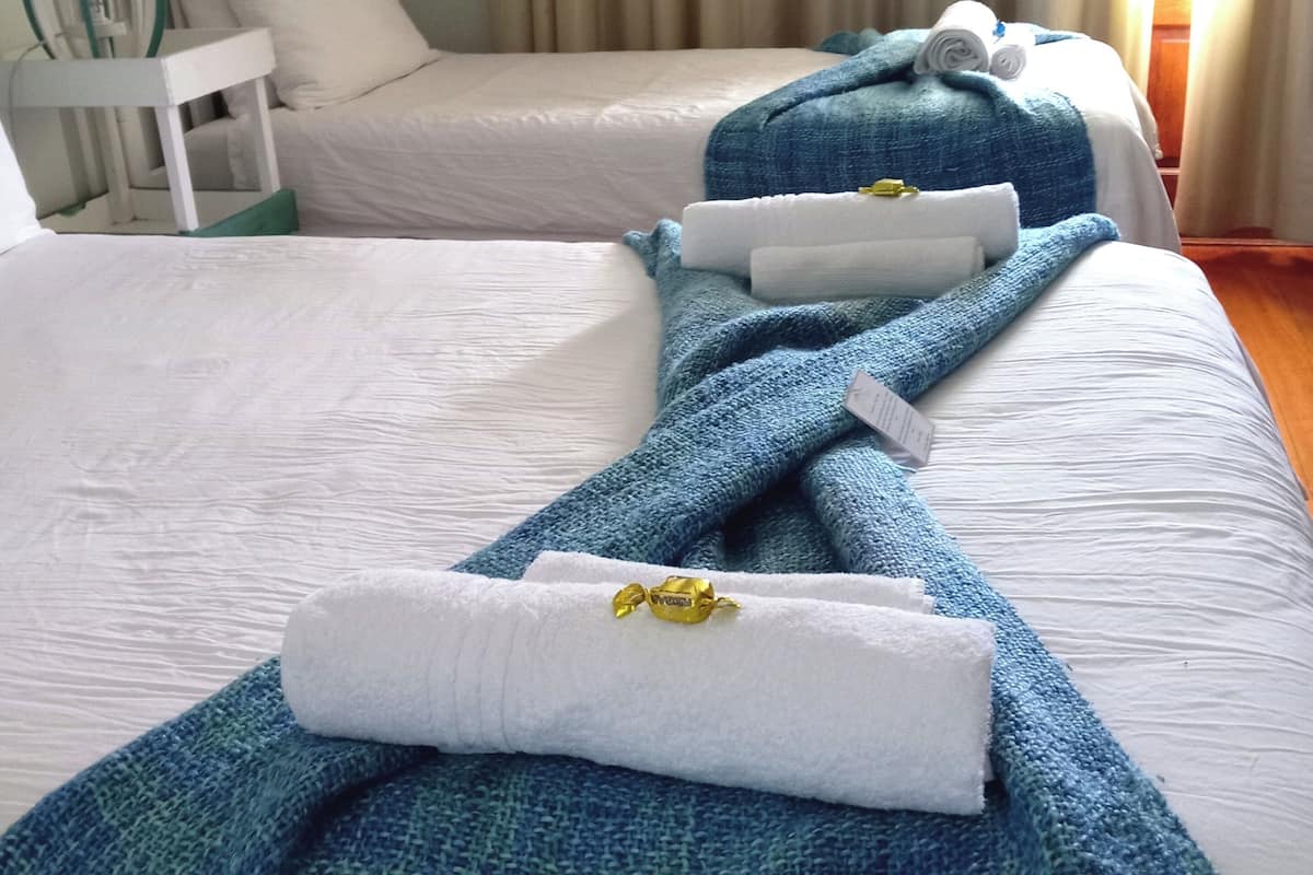 A hotel room with two beds, each with a white bedspread and blue blankets. There is a small table with a lamp and a chair.