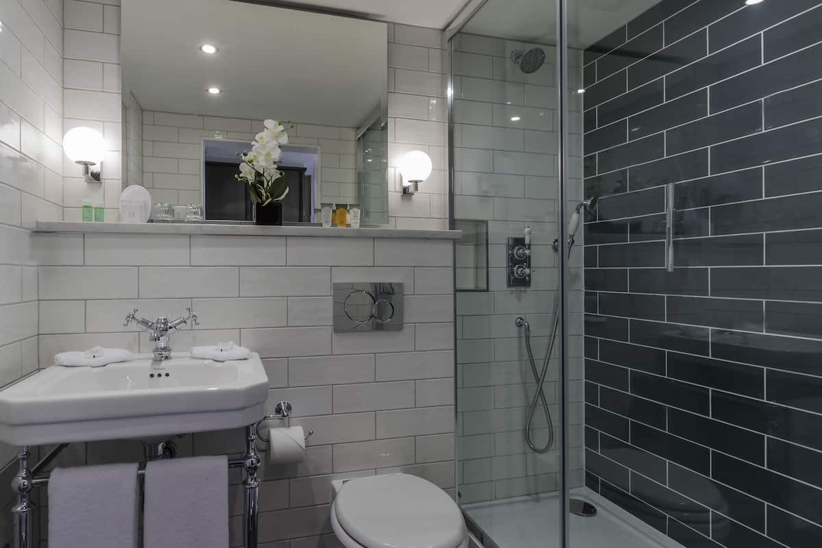 A modern bathroom with a white sink, toilet, and shower. There is a mirror above the sink and a shelf with toiletries.