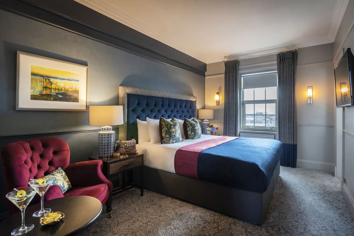 A hotel room with a large bed, a red armchair, a small table with two martini glasses, and a painting on the wall.