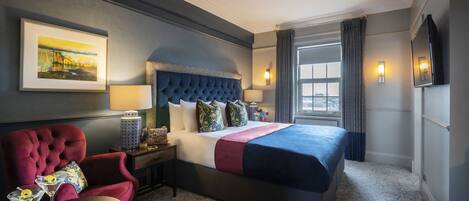 A hotel room with a large bed, a red armchair, a small table with two martini glasses, and a painting on the wall.