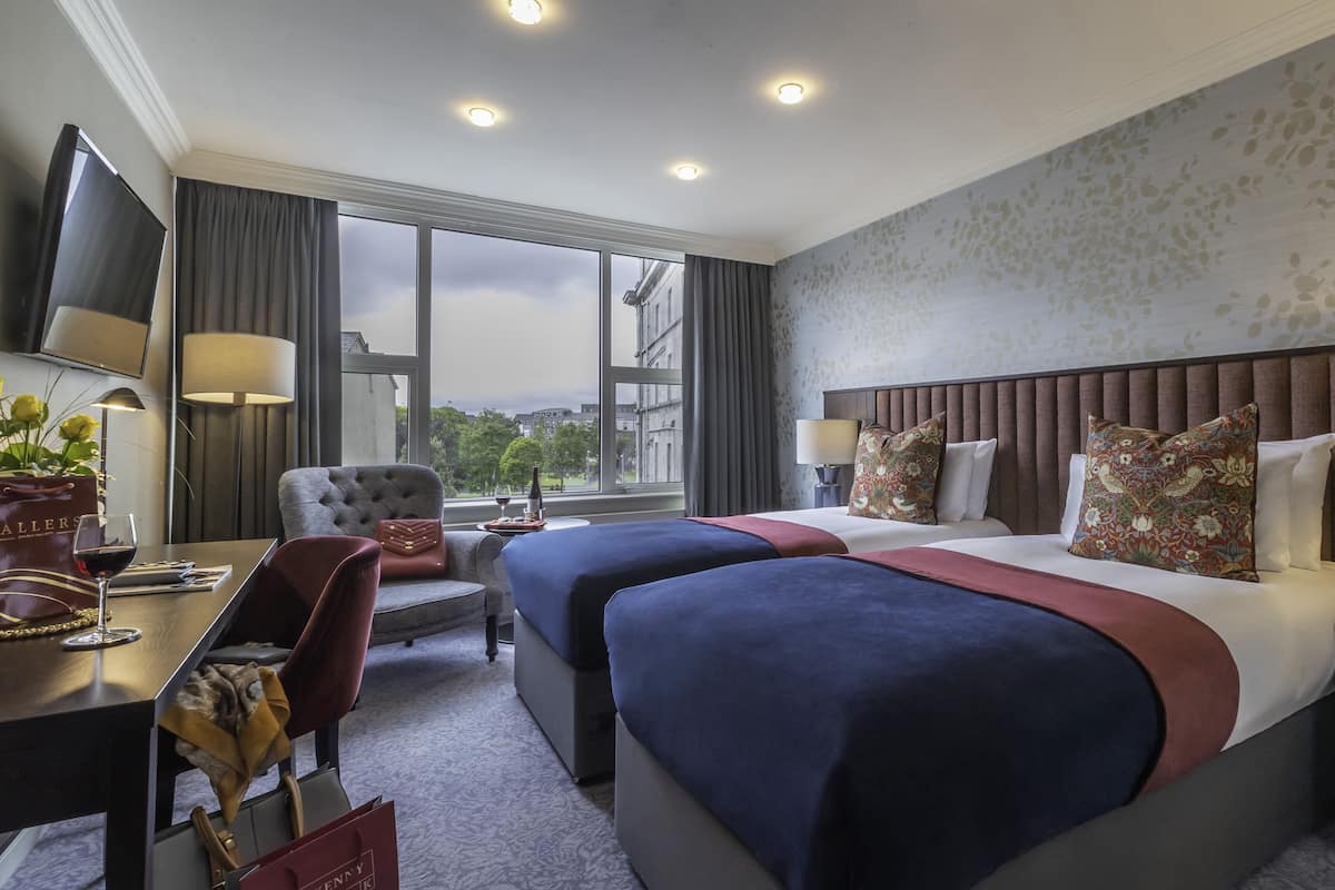 A hotel room with two beds, a desk with a chair, a TV, and a large window with a city view.