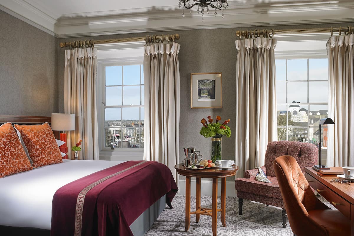 A hotel room with a large bed, a sitting area with a chair and table, a vase with flowers, and a view of the city through the window.