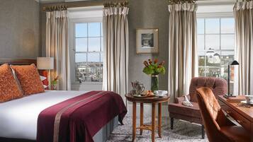 A hotel room with a large bed, a sitting area with a chair and table, a vase with flowers, and a view of the city through the window.