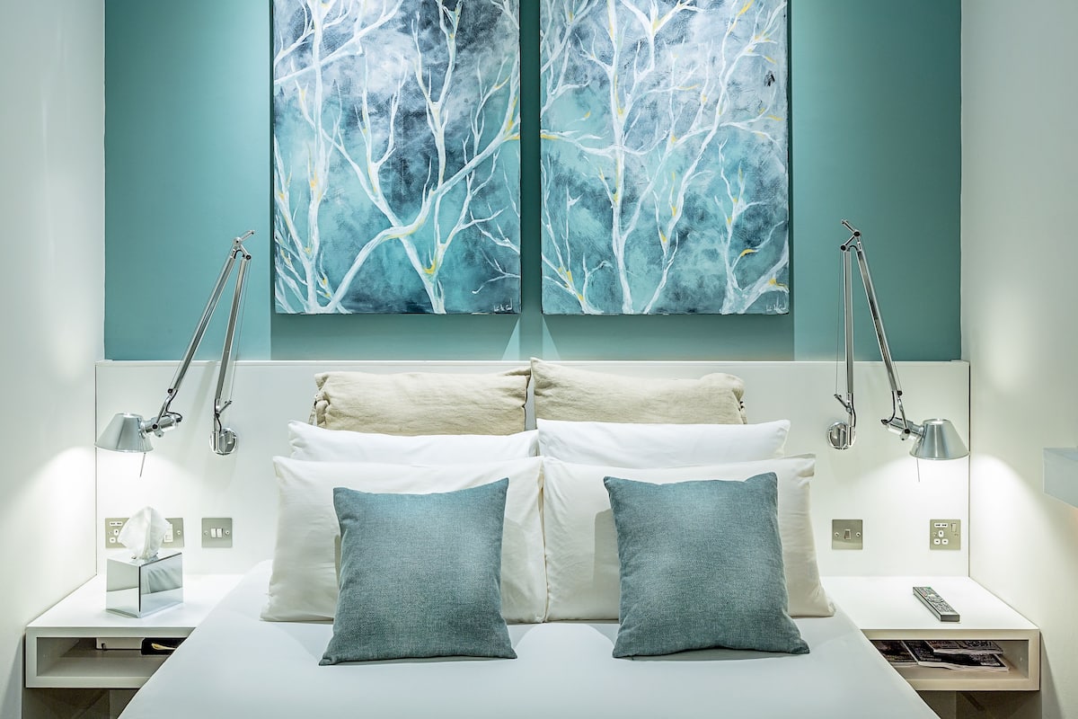 Une chambre à coucher moderne avec un lit, deux tables de chevet, deux lampes et deux tableaux abstraits accrochés au mur.