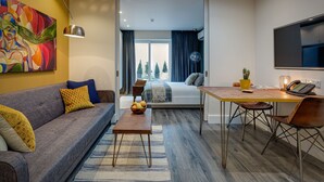 Una habitación de hotel moderna con sofá, mesa de centro y zona de comedor con sillas. Hay un cuadro en la pared, un televisor y una ventana con cortinas.