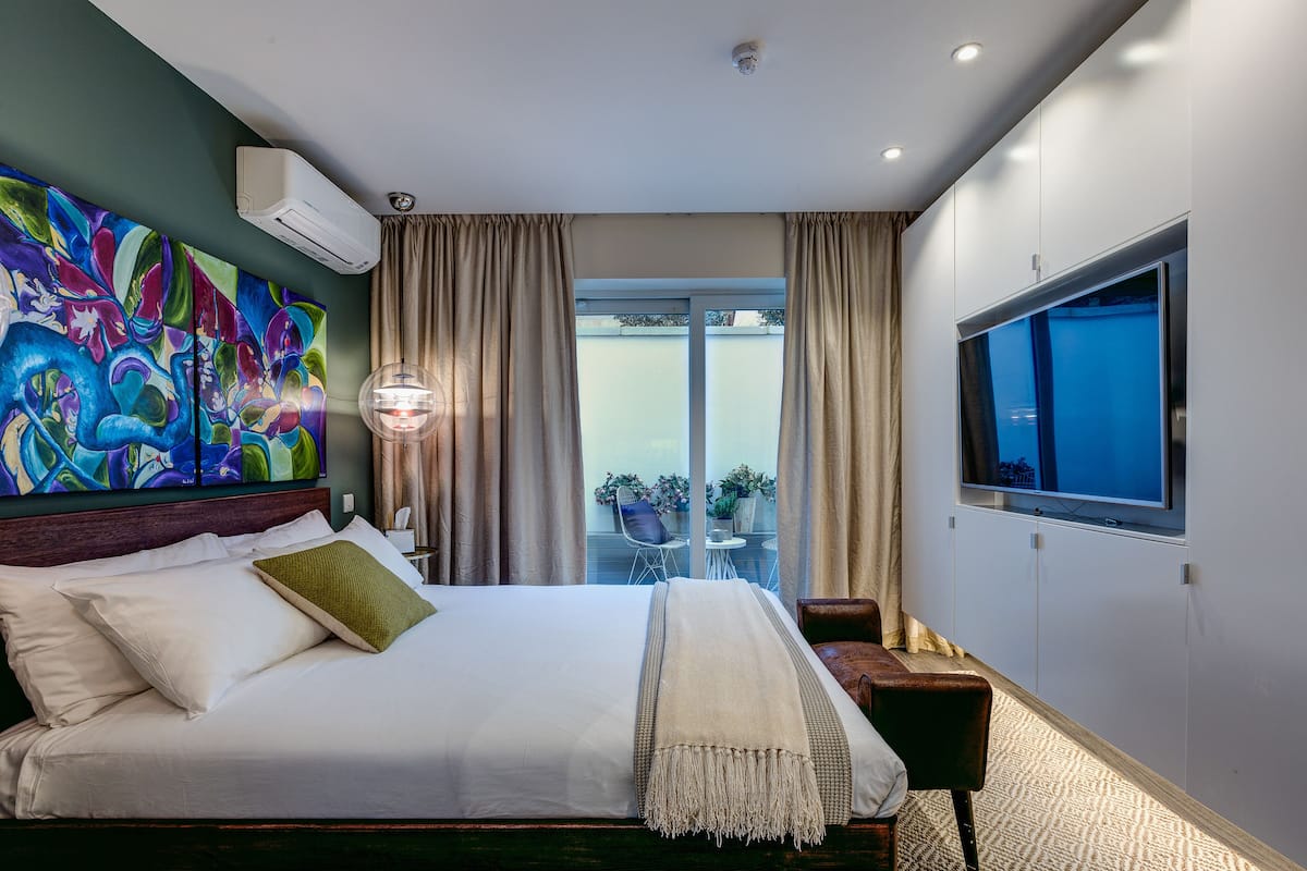 Une chambre d’hôtel avec un grand lit, un téléviseur fixé au mur, un balcon avec vue et une peinture abstraite aux couleurs vives accrochée au mur.