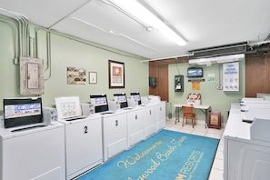 Eine Wäscherei mit nummerierten Waschmaschinen, einem blauen Teppich mit der Aufschrift "Welcome to Oakwood Beach Resorts" und einem an der Wand befestigten Fernseher.