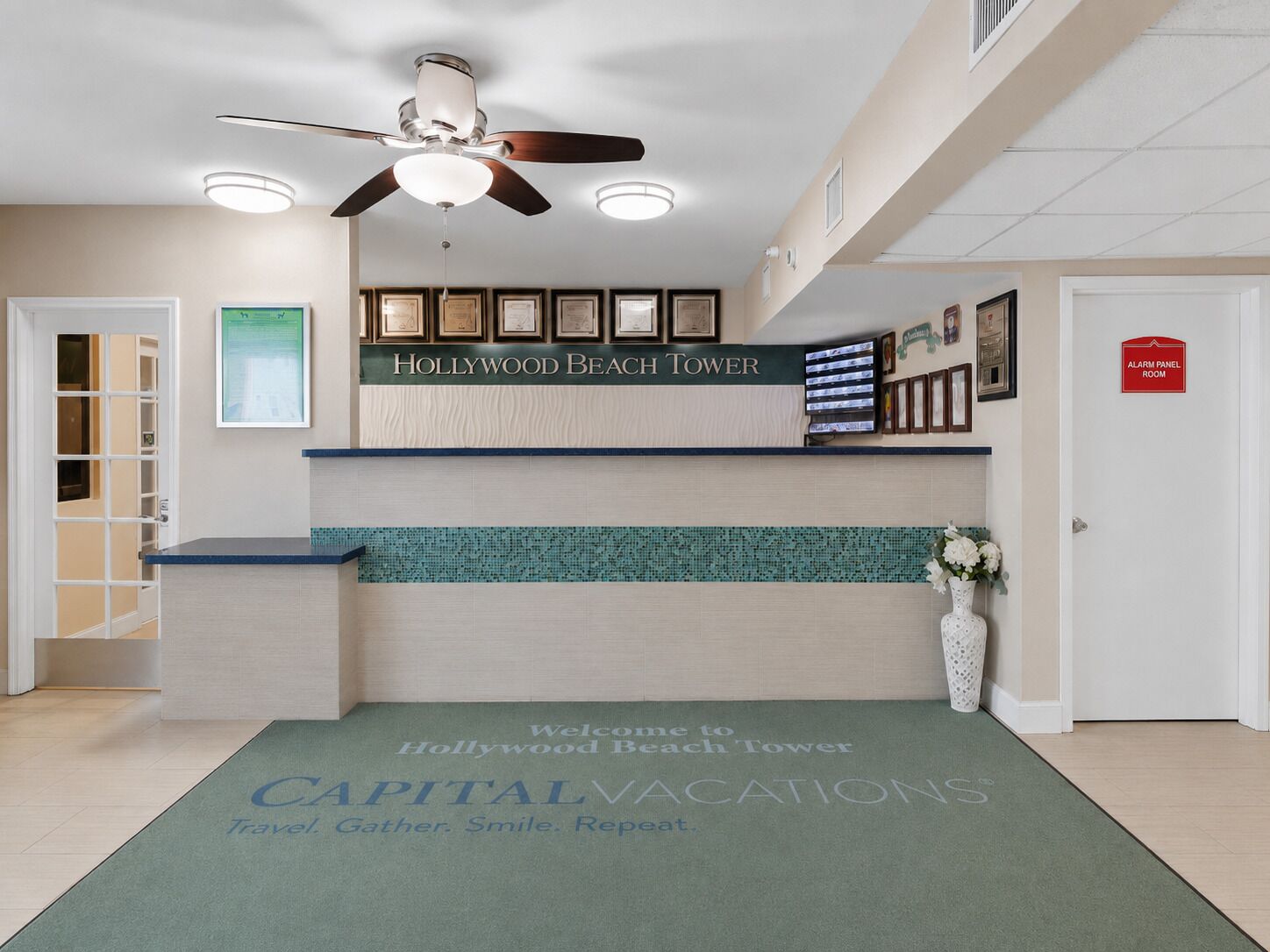 A reception area with a ceiling fan, framed pictures, and a welcome mat for Hollywood Beach Tower.