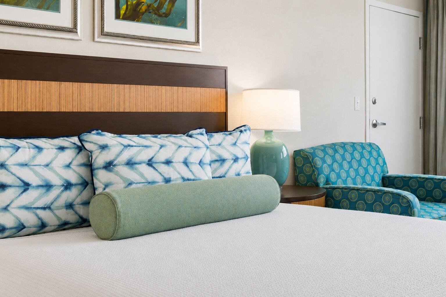 A hotel room with a bed, two pillows, a nightstand with a lamp, and two patterned chairs.