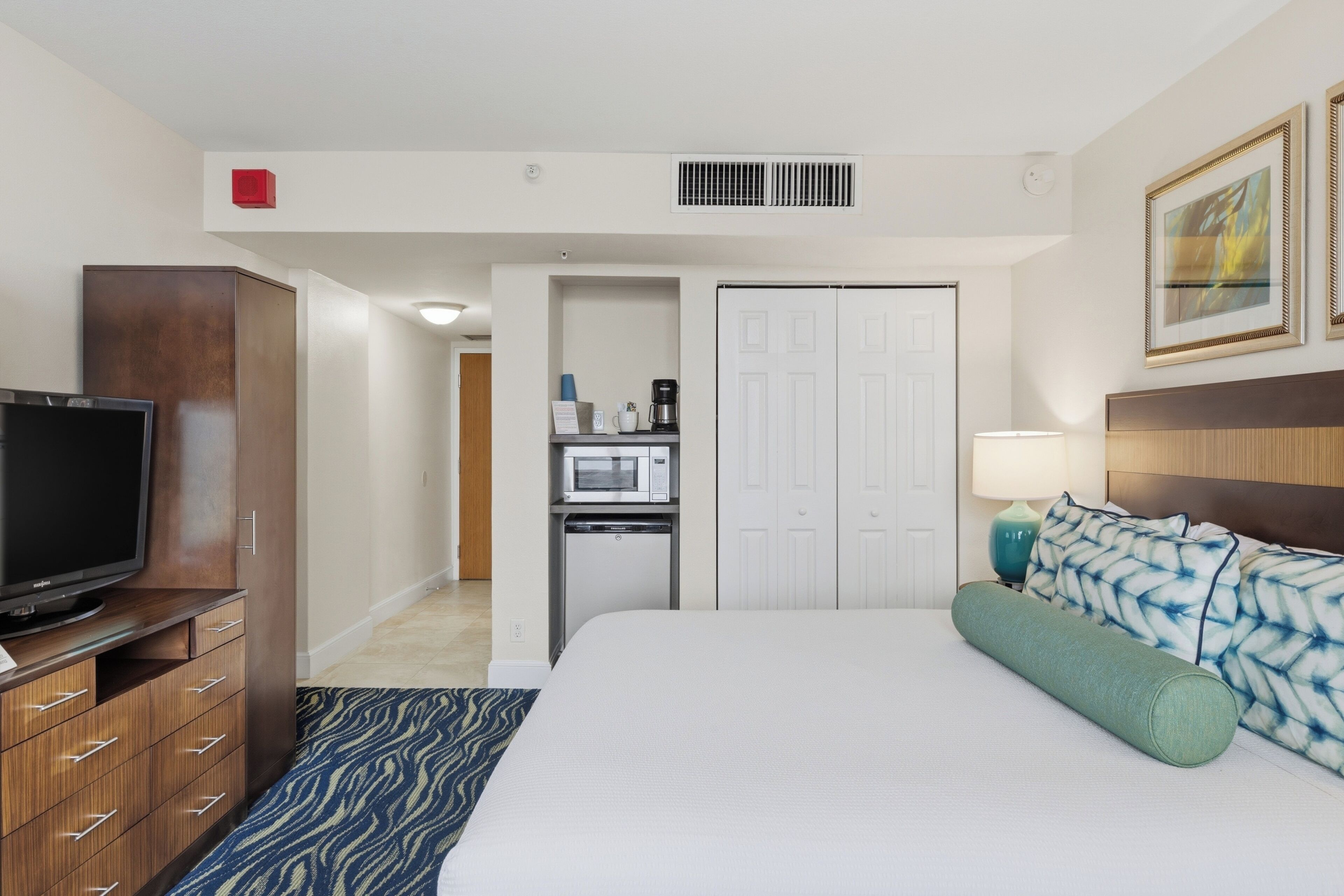 A hotel room with a flat-screen TV, a wooden dresser, a bed with patterned pillows, a microwave, and a small kitchenette.