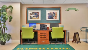 Een business center met twee bureaus, groene stoelen, een computer en lijstjes met kunstwerken aan de muur.