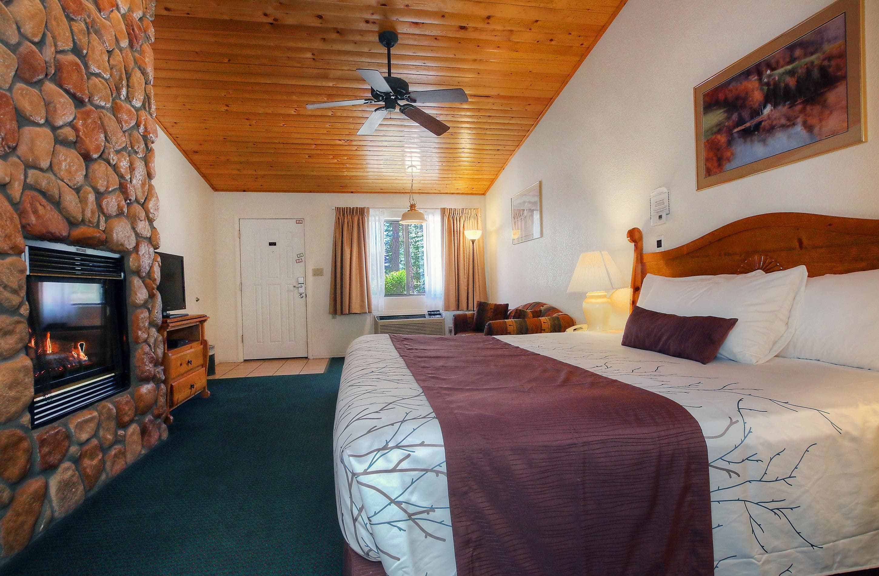 A bedroom with a stone wall, a fireplace, a bed with a headboard, a ceiling fan, and a window with curtains.