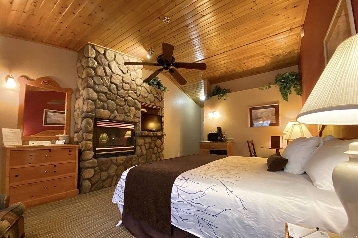 Un dormitorio con pared de piedra, techo de madera, cama con ropa de cama blanca y marrón, cómoda, televisor y ventilador de techo.