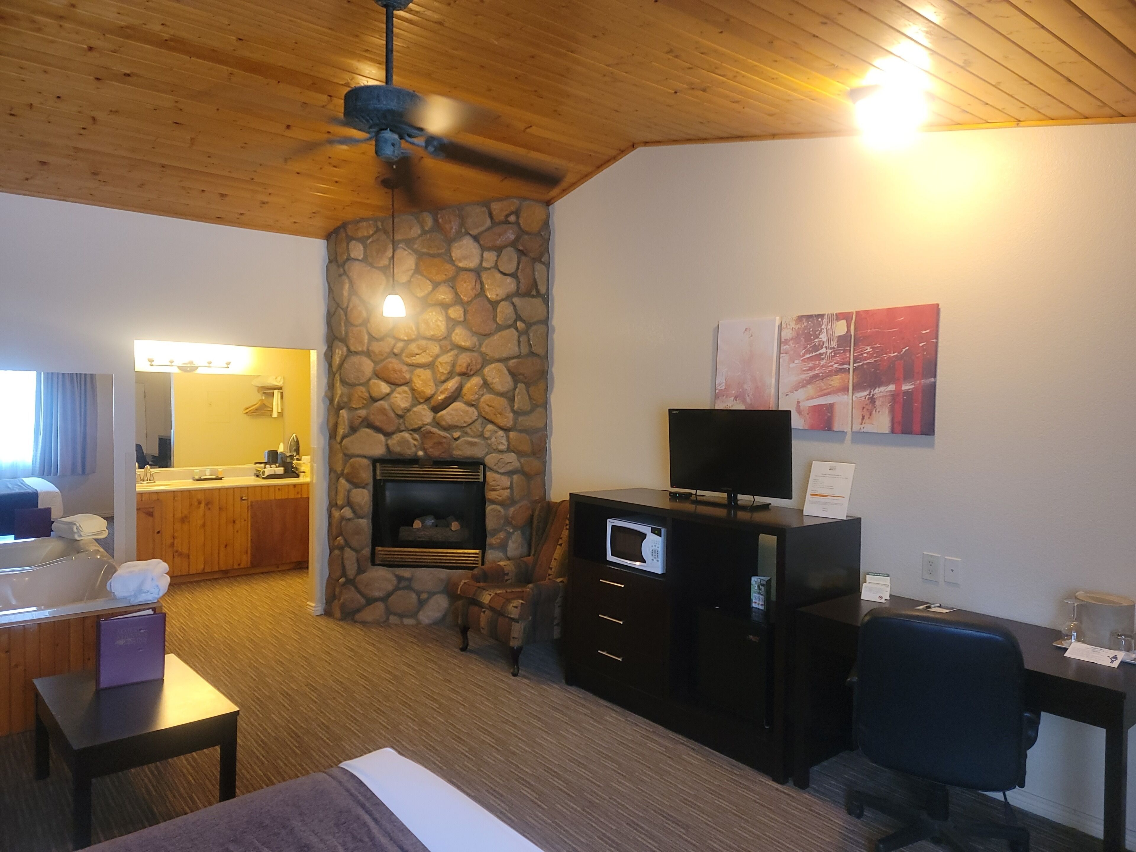A hotel room with a stone fireplace, a television on a cabinet, a desk with a computer, and a chair.