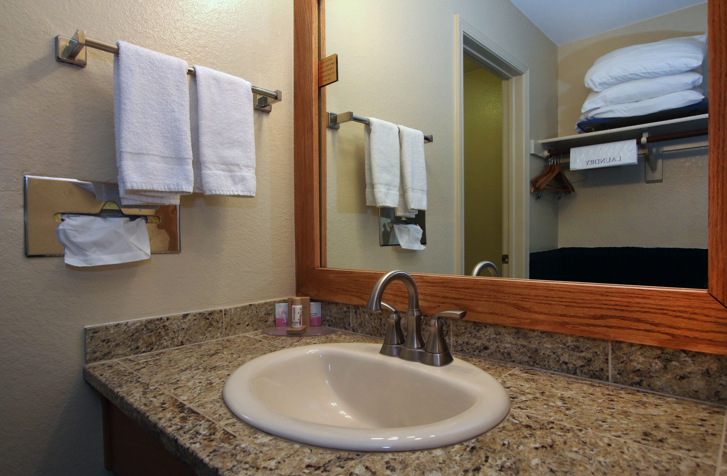 A bathroom with a granite countertop, a white sink, and a mirror. There are towels hanging on a rack and a shelf with folded towels.