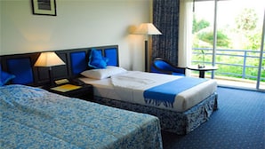 A hotel room with two beds, a nightstand, a lamp, a chair, and a view of greenery outside. - Orient D'angkor (Siem Reap)