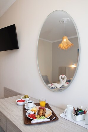 A breakfast tray with food and drinks on a wooden counter, a mirror reflecting a swan-shaped towel, and a television mounted on the wall.