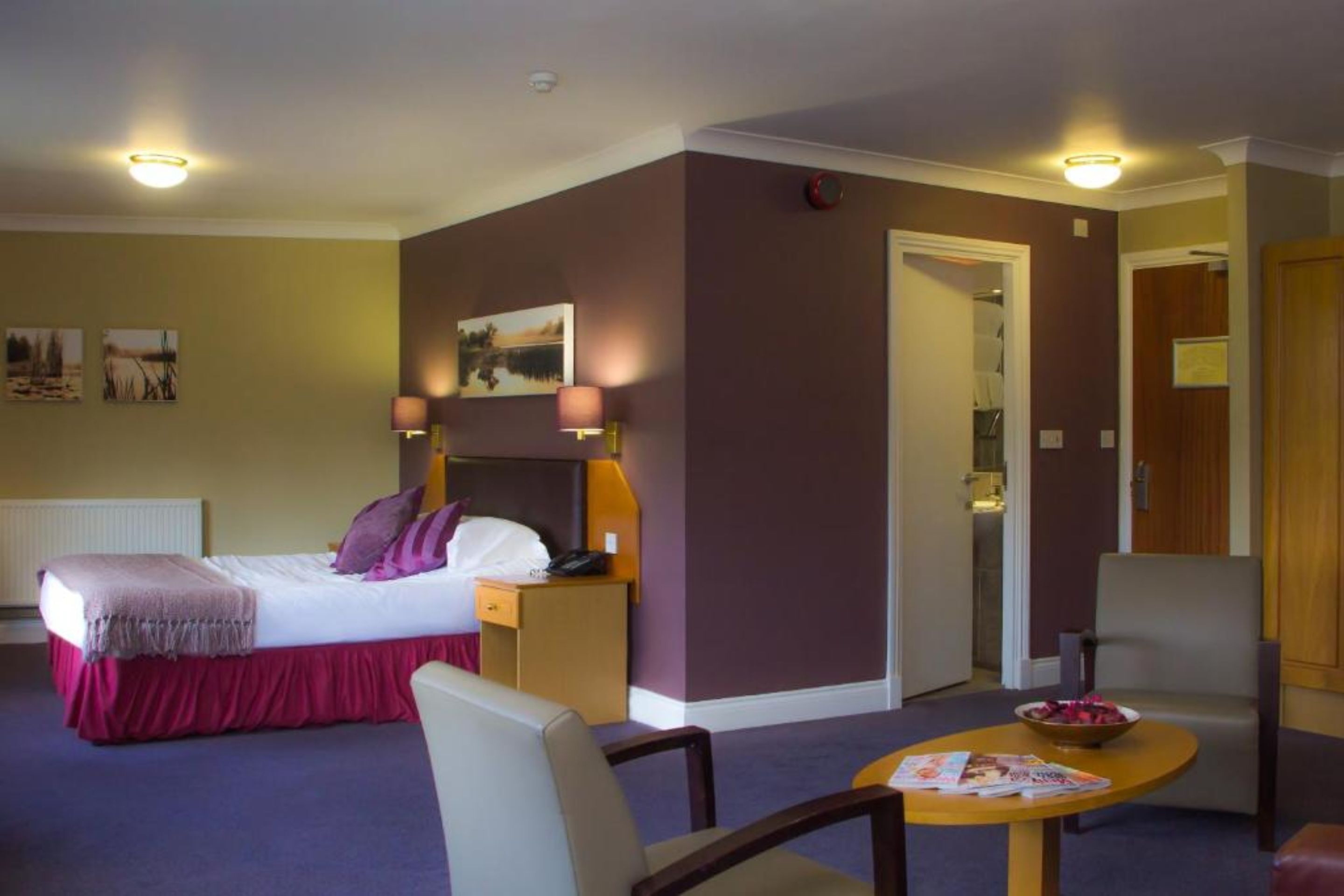 A hotel room with a bed, bedside table, lamp, chair, and a small table with a bowl and magazine.
