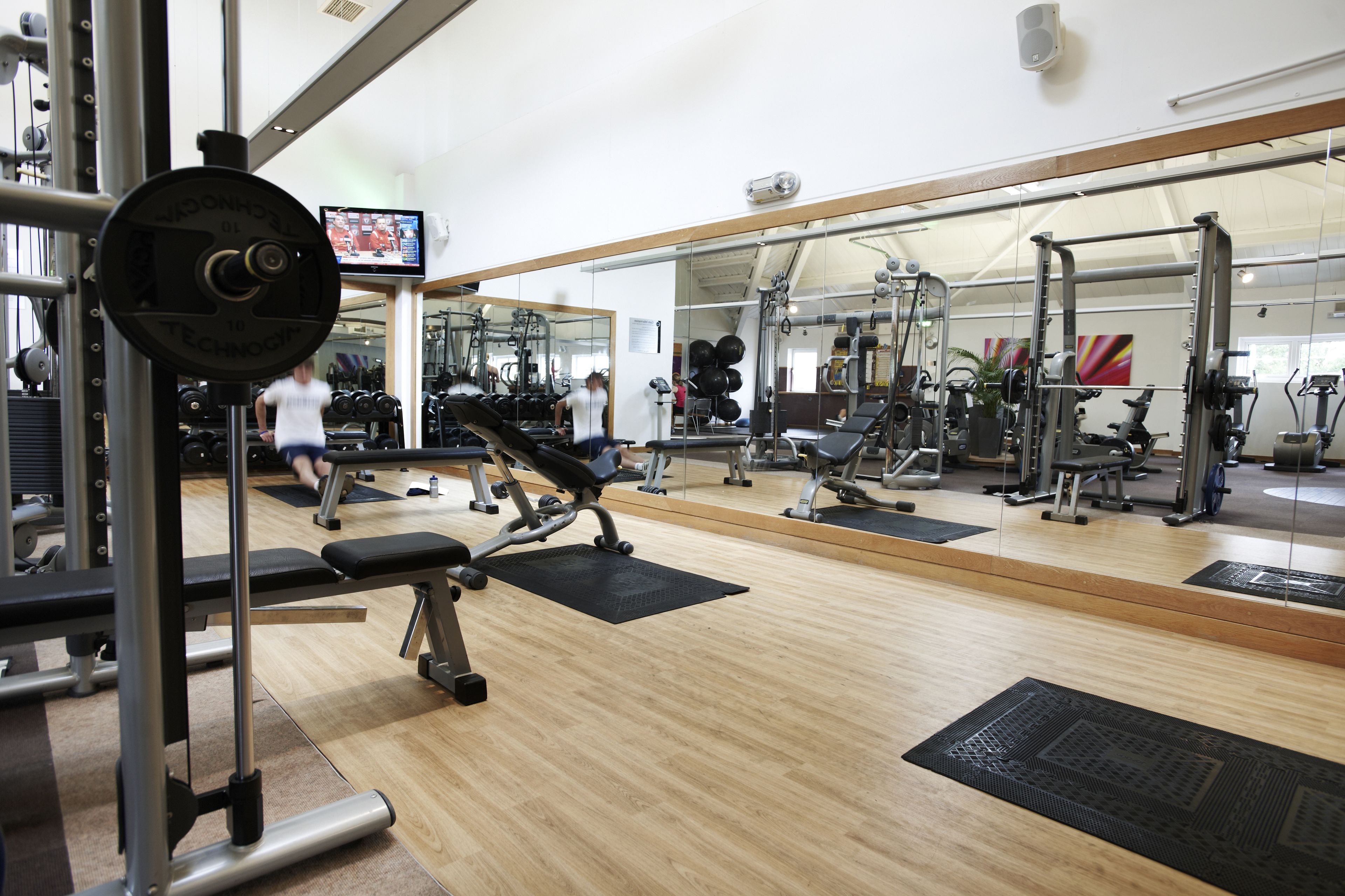 Un gimnasio con equipamiento de levantamiento de pesas, espejos y una pantalla de televisión.