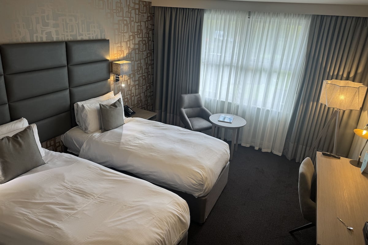 Une chambre d’hôtel avec deux lits, un bureau, une chaise, une petite table et une fenêtre avec des rideaux.