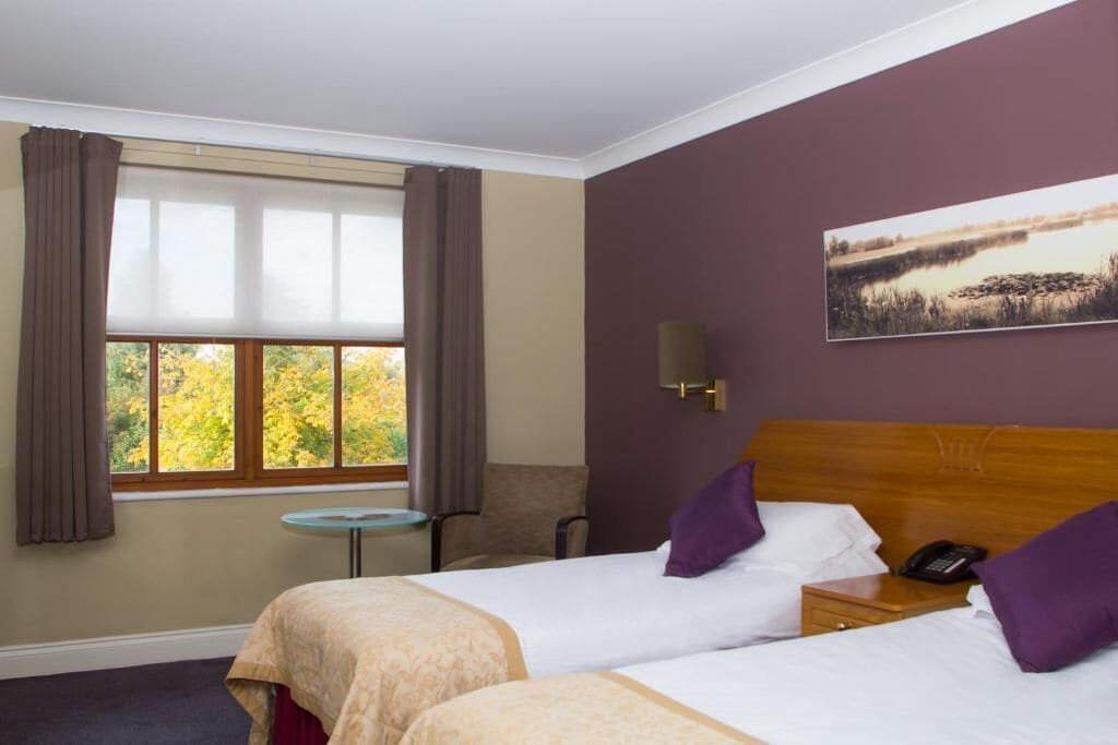 Ein Hotelzimmer mit zwei Betten, einem kleinen Tisch, einem Stuhl, einem Nachttisch mit Telefon und einem Fenster mit Vorhängen.