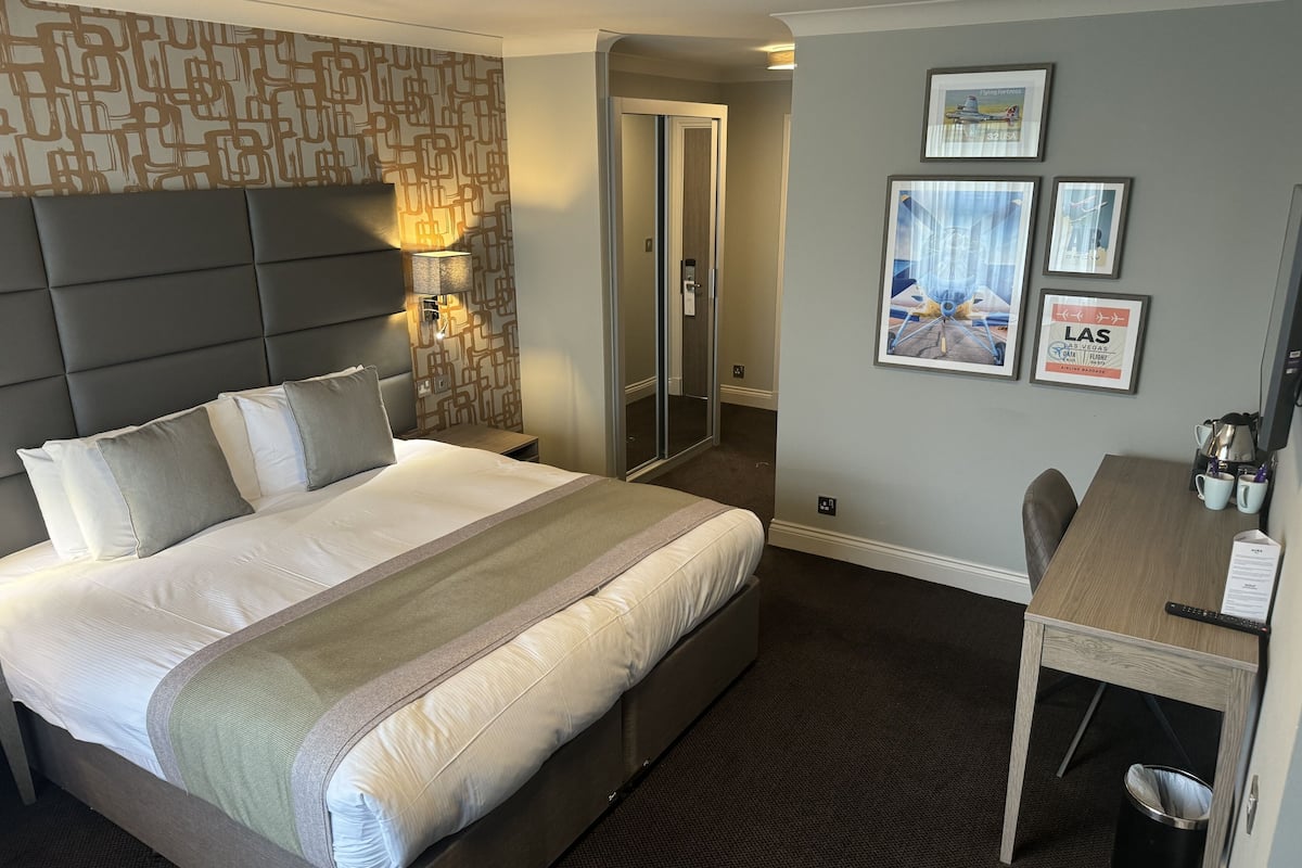 Une chambre d’hôtel avec un grand lit, un bureau et une chaise. Les murs sont ornés de tableaux encadrés et un miroir est placé au-dessus du lit.