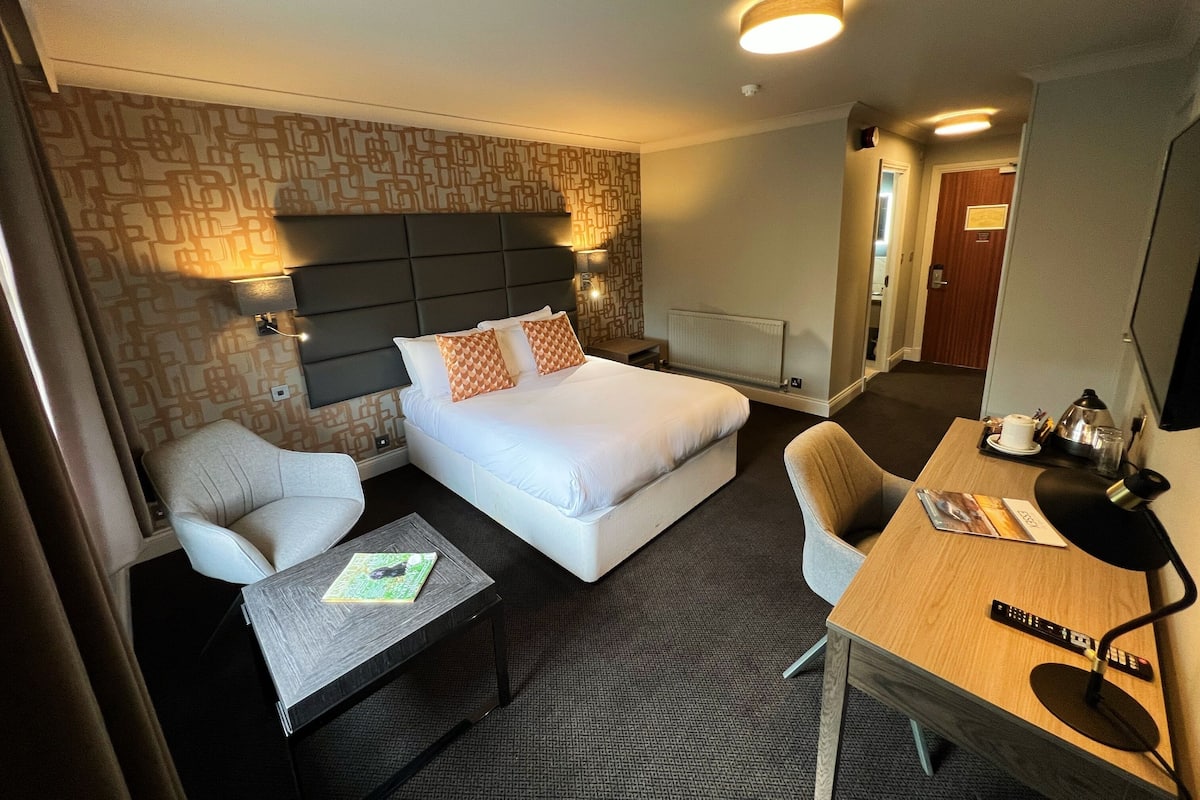 Une chambre d’hôtel comprenant un lit, deux fauteuils, une petite table, un bureau et un téléviseur.