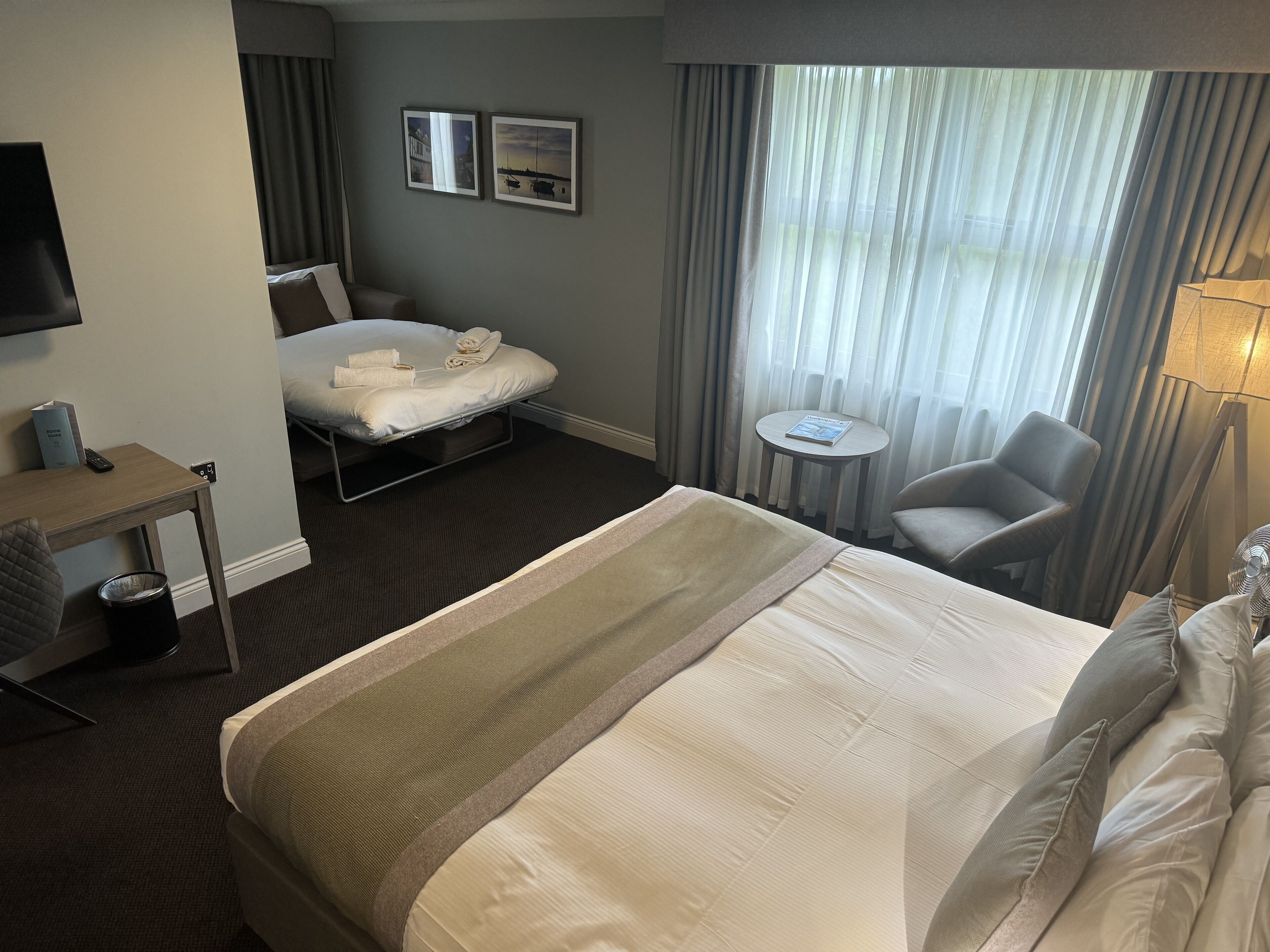 A hotel room with a large bed, a desk, a chair, a small table, and a TV.