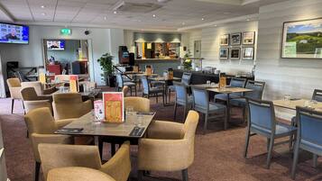 A hotel dining area with tables and chairs, a kitchen in the background, and a TV screen displaying a person.
