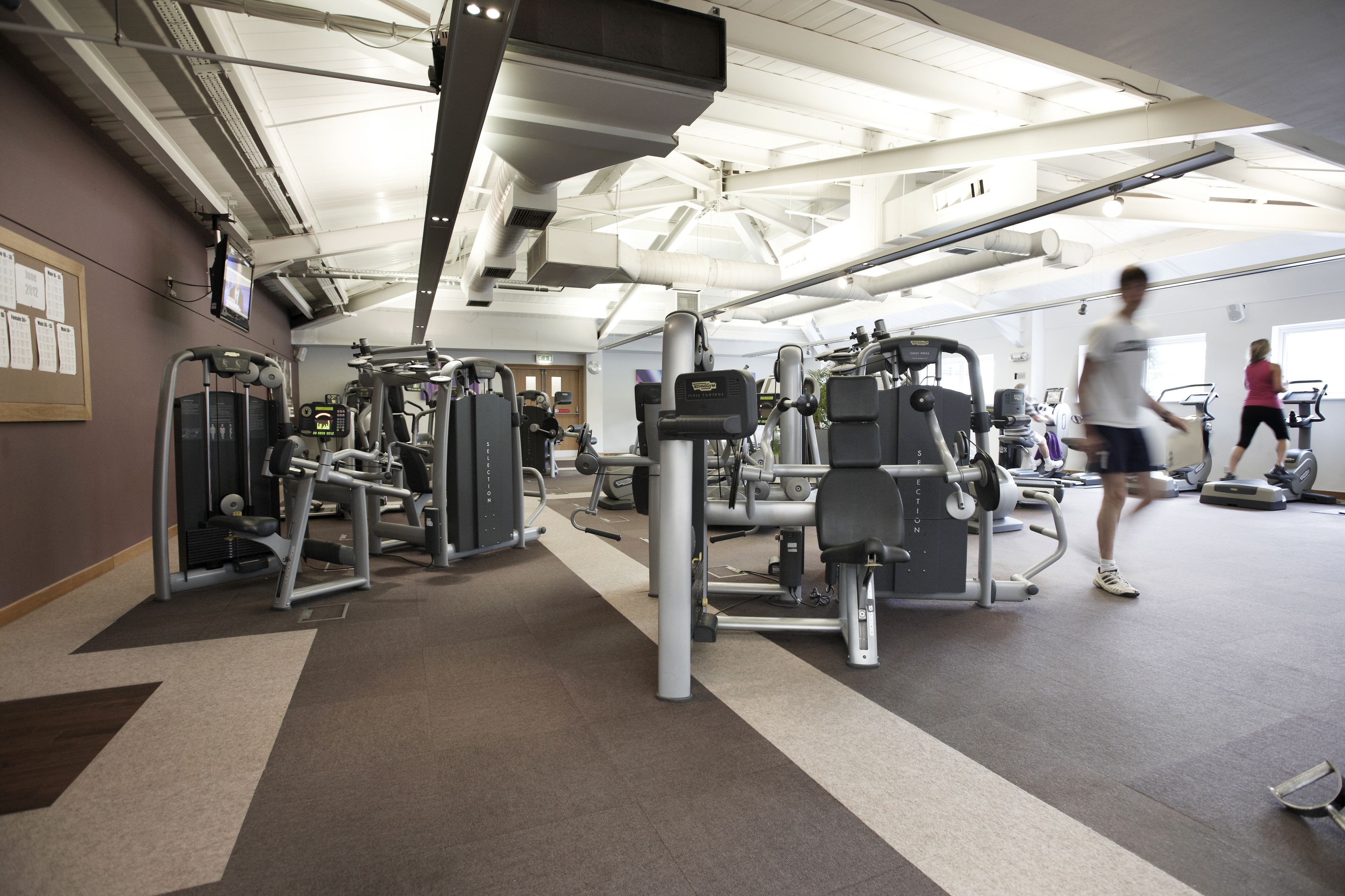 Un gimnasio con diversas máquinas de ejercicio, un espejo grande y un techo con vigas y luminarias a la vista.