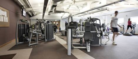 A gym with various exercise machines, a large mirror, and a ceiling with exposed beams and lighting fixtures.
