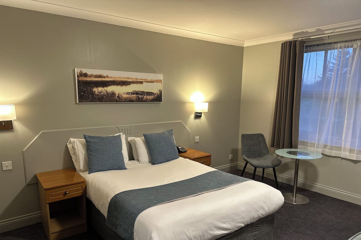 Une chambre d’hôtel avec un lit, des tables de chevet, une chaise, une petite table, un tableau au mur et une fenêtre avec des rideaux.
