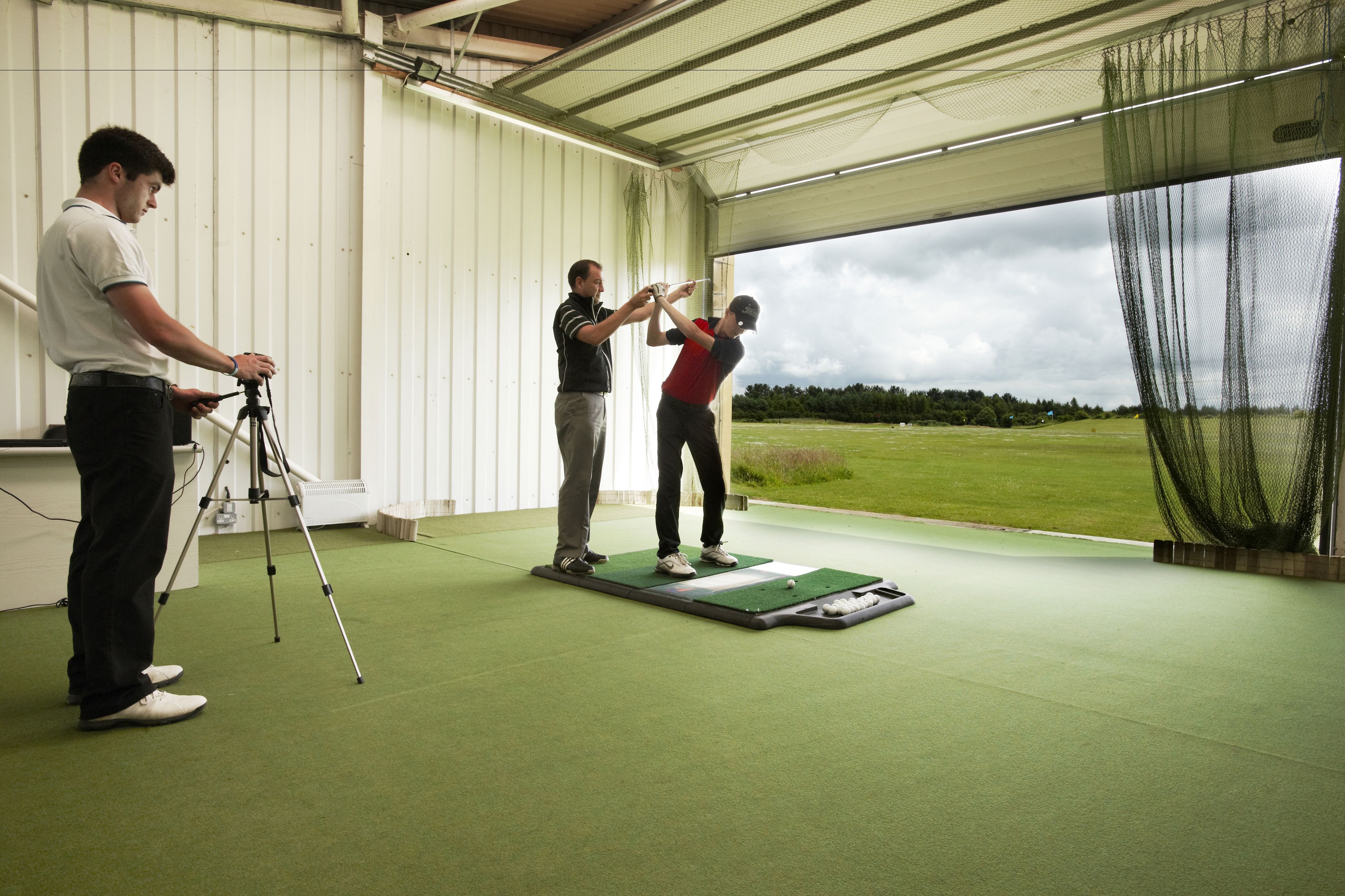 A golf training facility with a green practice area, a netted practice area, and a view of the outdoors.