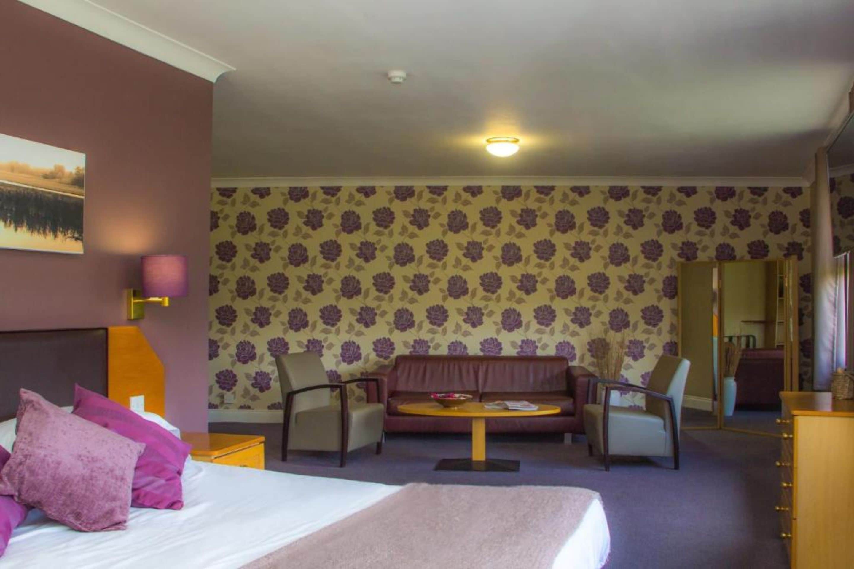 Habitación de hotel con cama, sofá, dos sillones, una mesita y una pared con papel tapiz floral.