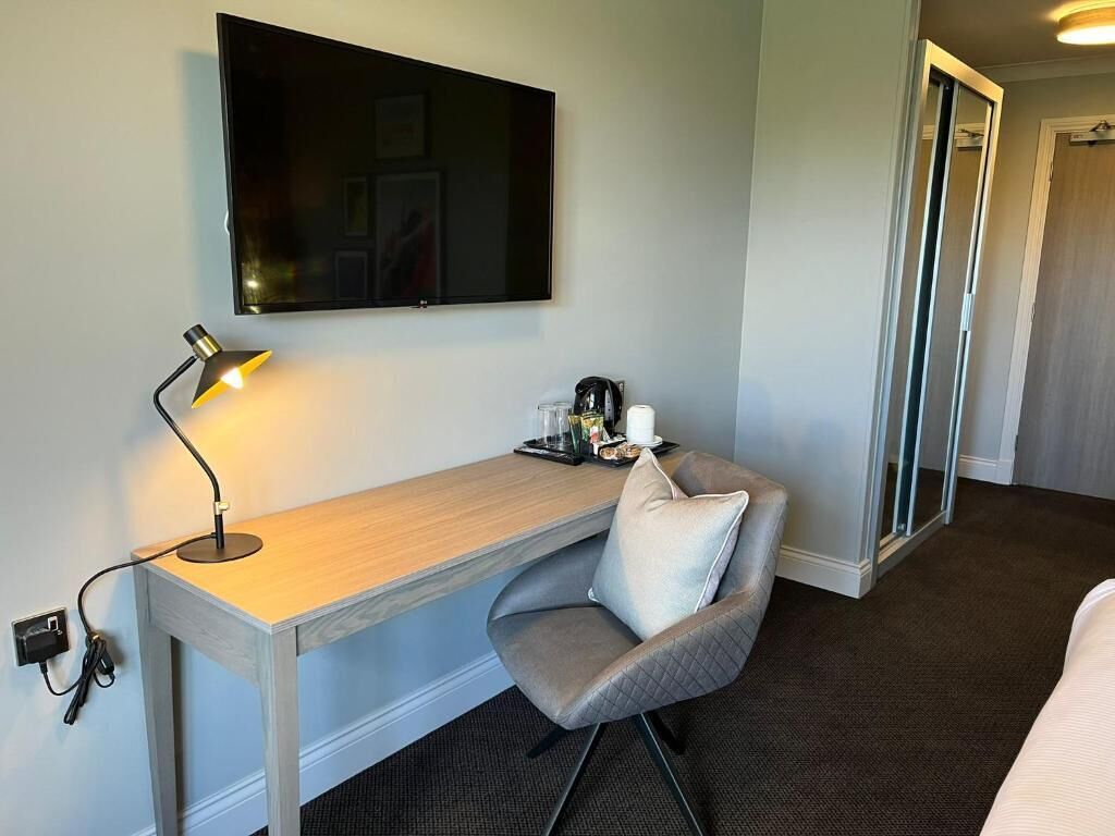 A hotel room with a desk, chair, flat-screen TV, bedside table, and a bed.